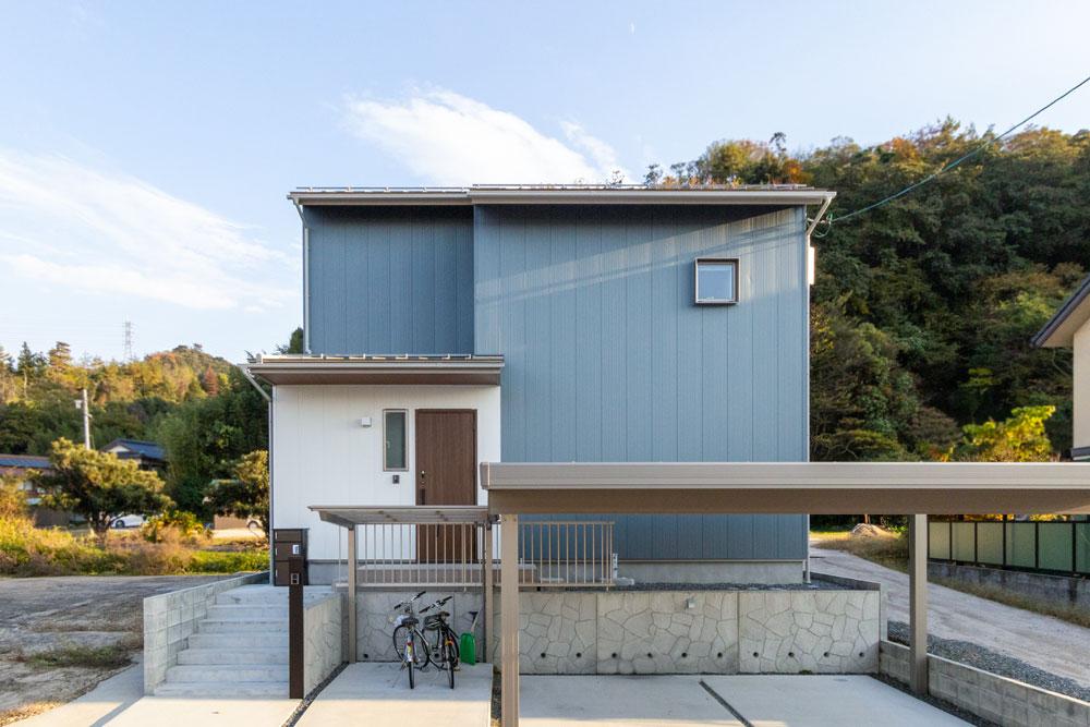 ブルーグレーの金属サイディングに白い壁。木目の玄関扉がアクセント。